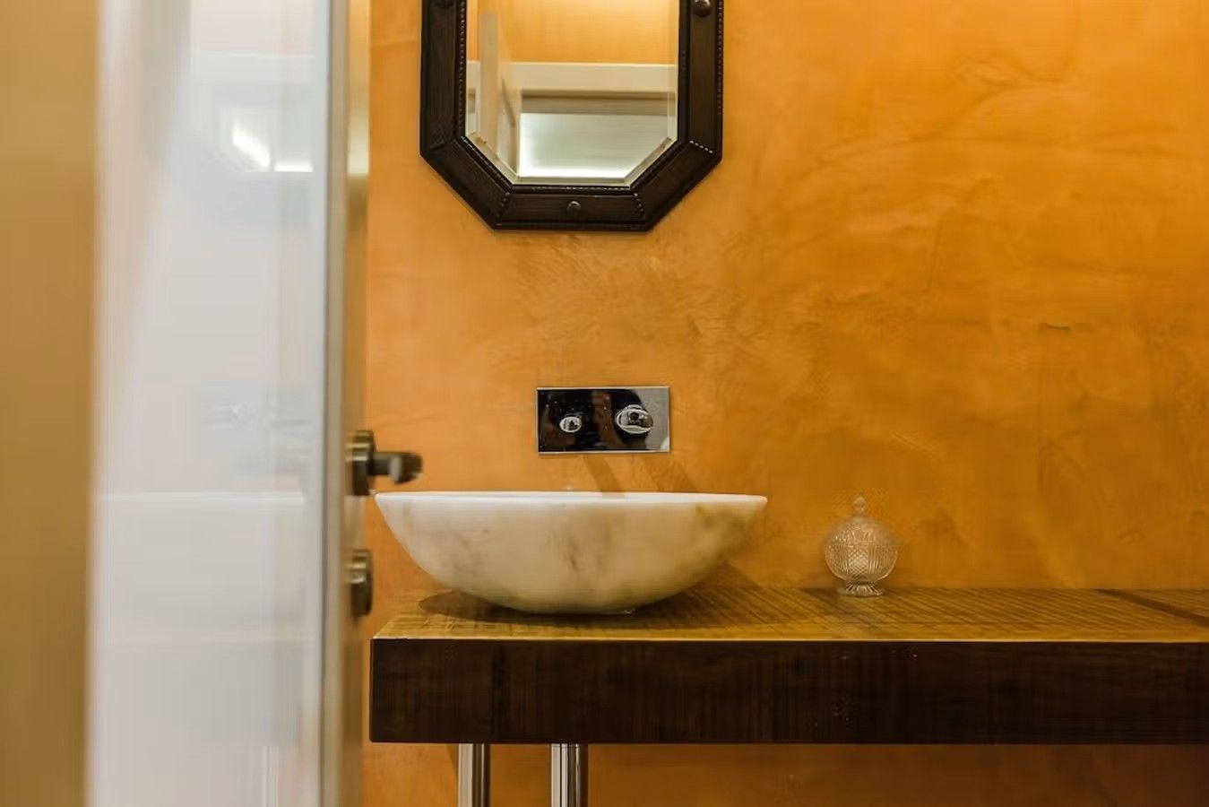 Fra et baderom designet med en rustikk og orange vegg, et mindre speil på veggen og et vaskevannsfat i marmor.