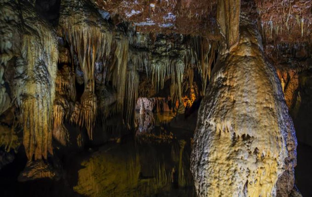 Inne i en dryppsteinsgrotte med stalaktitter og stalagmitter omgitt av vann og opplyst av kunstig lys.
