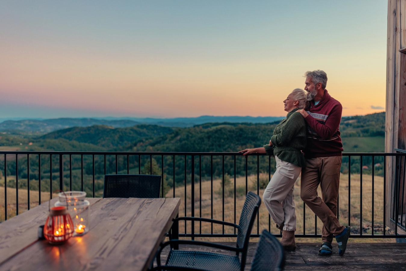Et eldre par står på balkongen under en solnedgang og nyter utsikten over et grønnkledd fjellandskap