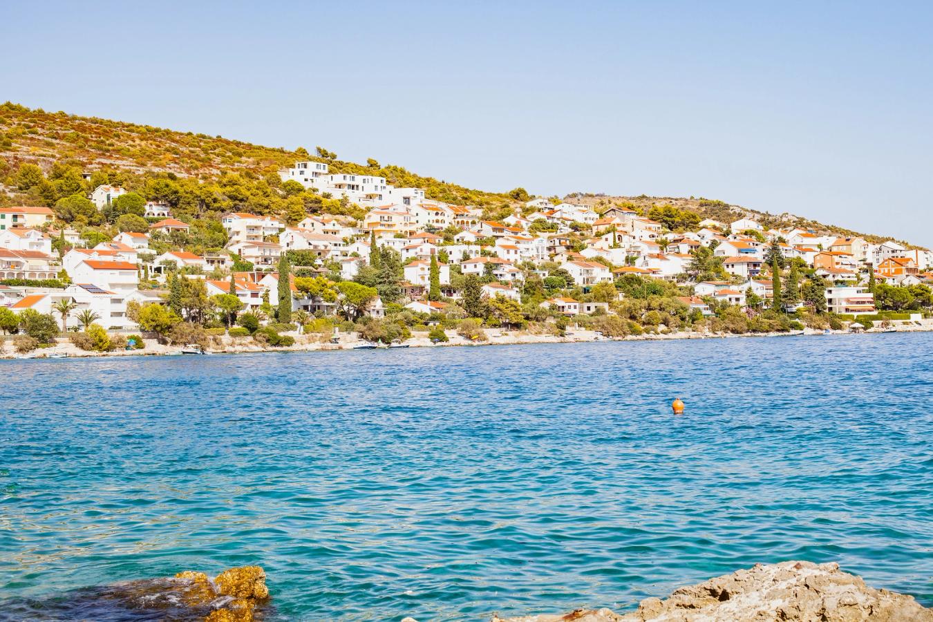 Udsigt over en charmerende kystby i Kroatien med hvide huse og orange tegltage, som ligger spredt op ad en grønklædt bakke. Foran byen ses det klare, turkisblå Adriaterhav, og kystlinjen er omgivet af klipper og små både. Himlen er skyfri og lyseblå, hvilket fremhæver den solrige middelhavsstemning.