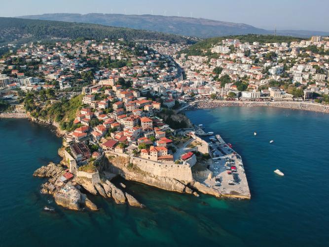 Bugten ved Ulcinj i Montenegro. Den gamle bydel med røde tegltag strækker sig ud over vandet, der er turkisblåt og klart. På vandet ses en lille båd og i baggrunden strækker resten af byen sig indover landskabet.