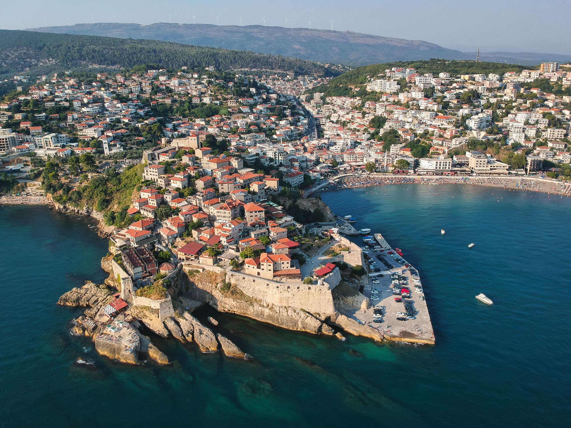 Bukta ved Ulcinj i Montenegro. Den gamle bydelen med røde takstein strekker seg ut over det turkisblå og klare vannet. På vannet sees en liten båt, og i bakgrunnen strekker resten av byen seg inn over landskapet.