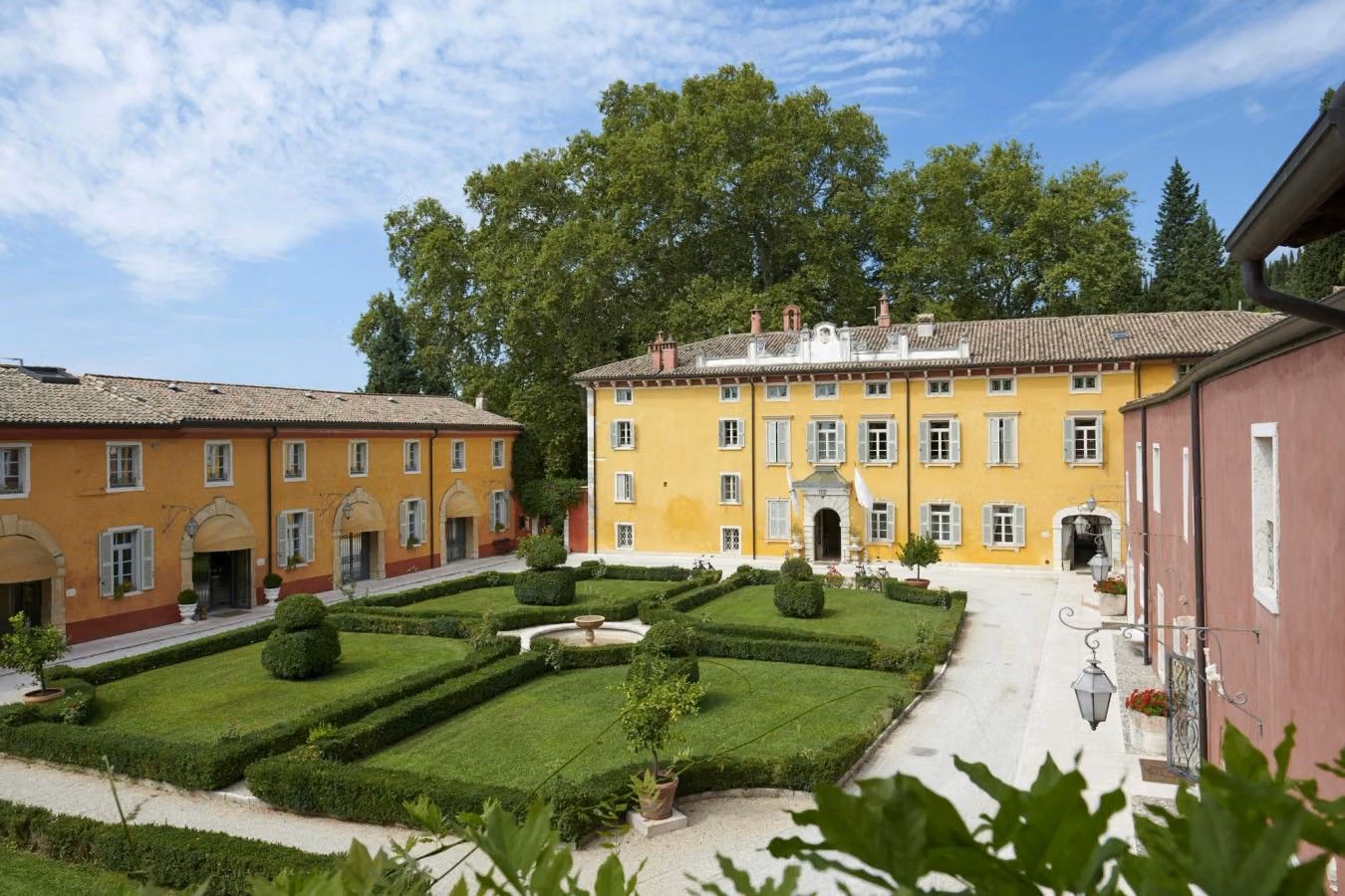 En venetiansk villa fra 1500-tallet i okkergul farve omkranser en mindre, grøn have. Store egetræer i baggrunden, alt under en blå himmel.