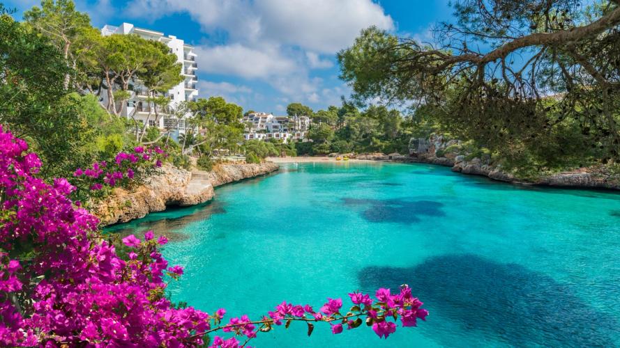 Her ses Cala d’Or-bugten ved byen Cala d’Or på Mallorca. Turkisblåt vand inde i bugten, som ender i en sandstrand. Farverig og frodig vegetation langs bugtens sider.