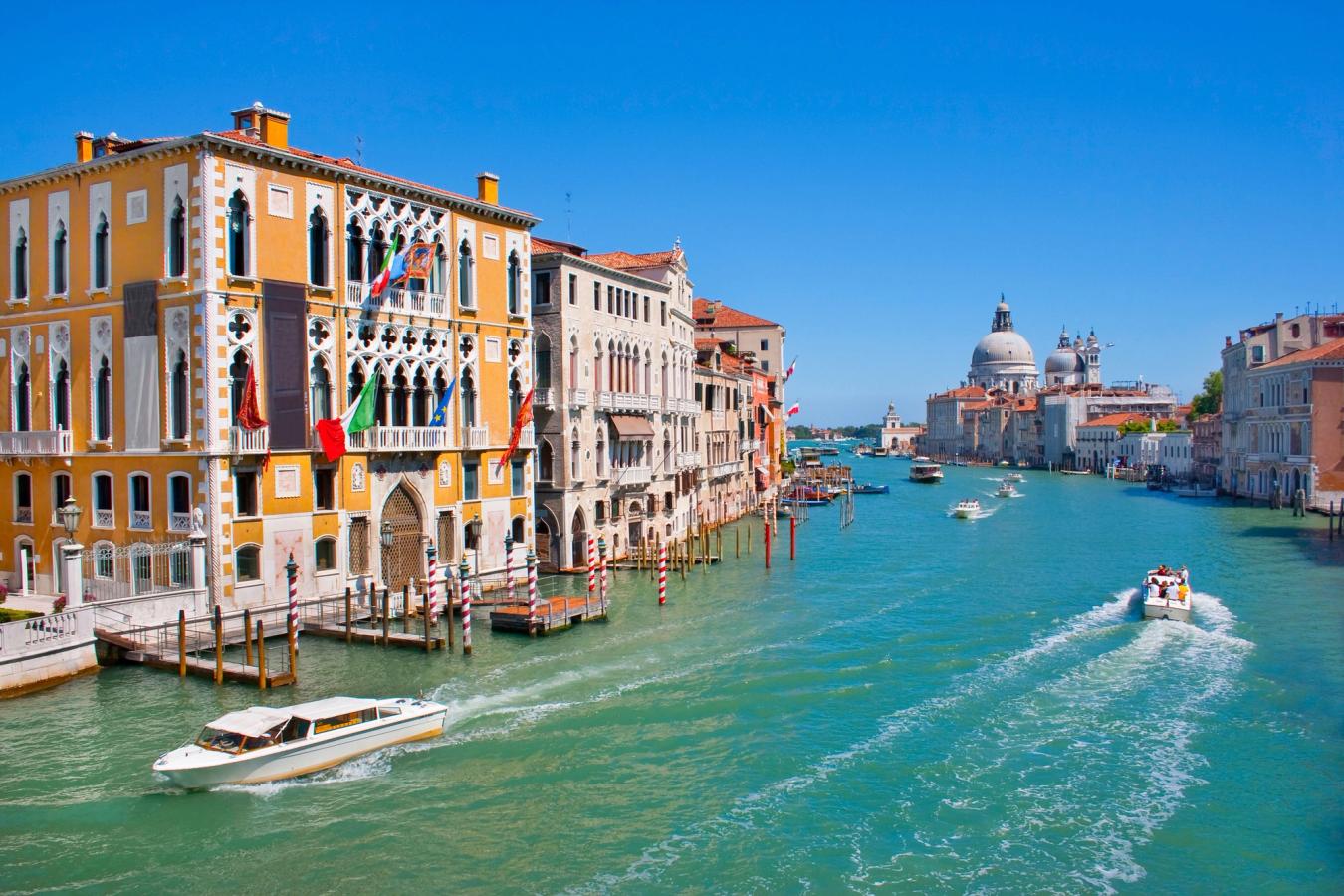 Bildet viser det turkise vannet i Canal Grande i Venezia. Noen båter flytter seg på vannet under en skyfri himmel, mens vi ser den klassiske arkitekturen
