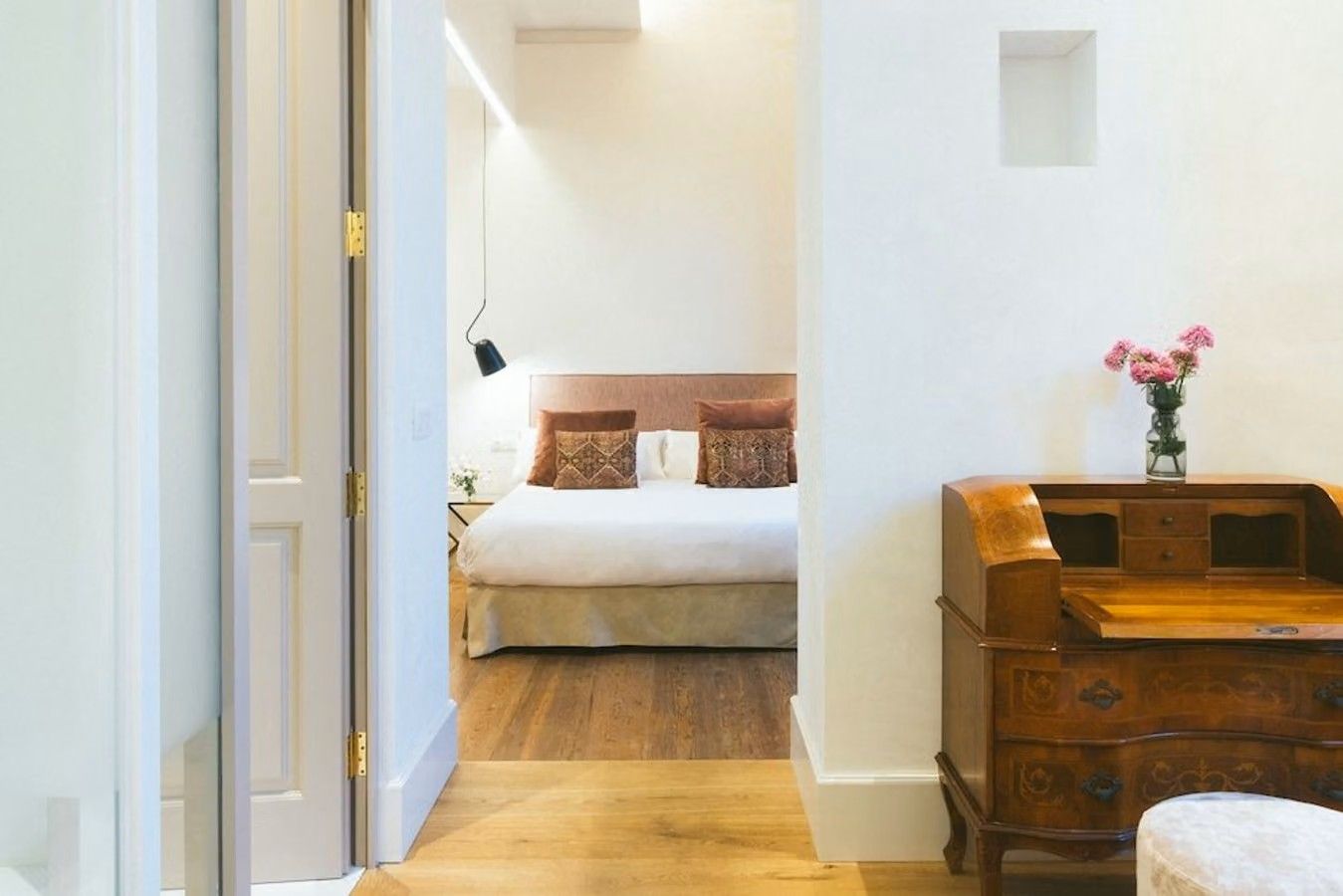 hotellet Veintiuno Emblematic Hotels By Airnest i Las Palmas på Gran Canaria. Rommene er hvitkledde. I det nærmeste rommet ser vi et skrivebord i treverk på høyre side, hvor det så er en åpen inngang til soverommet. Der ser vi en hvit dobbeltseng med røde puter på.