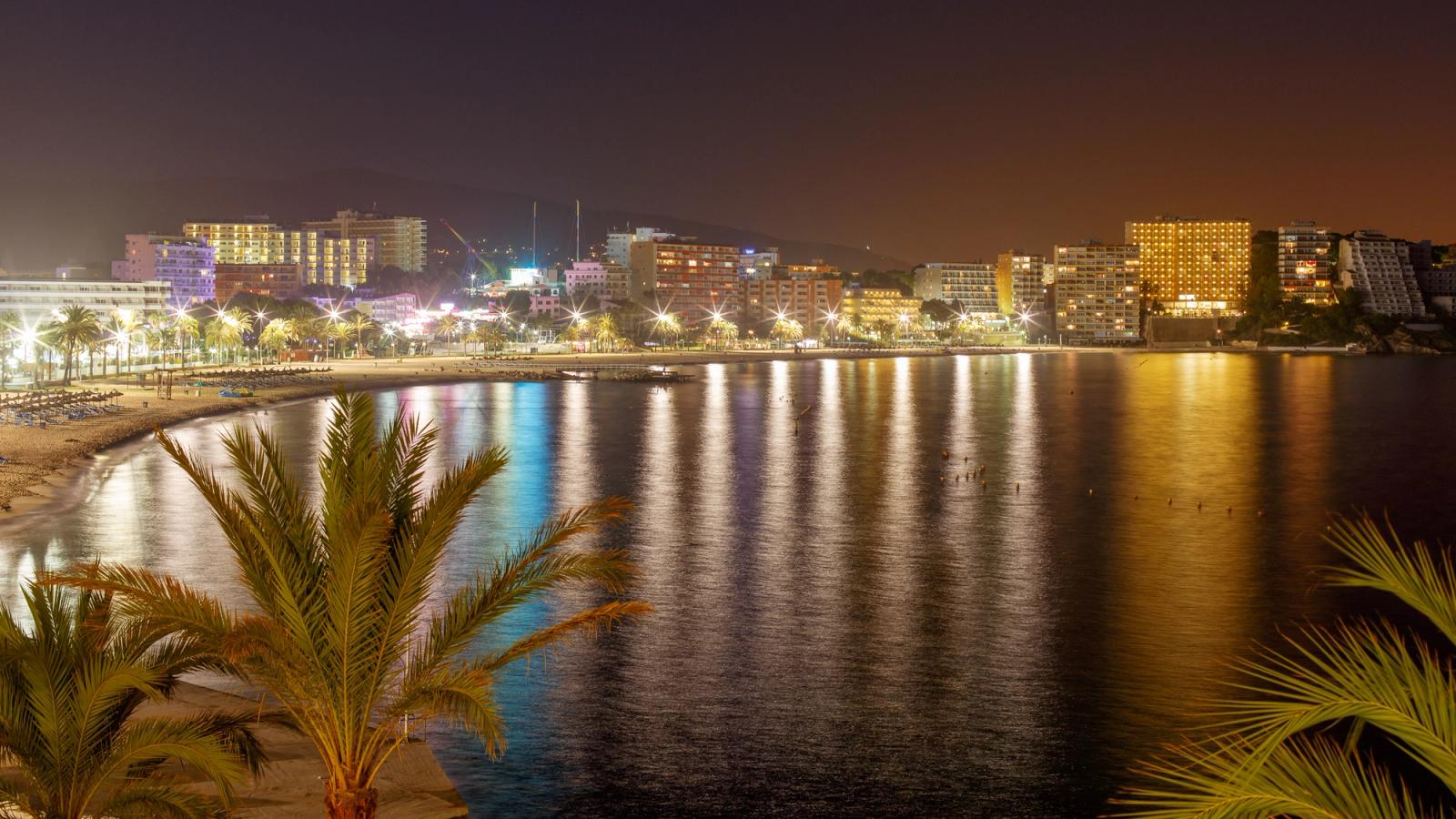 Bilden visar Magaluf och huvudstranden på kvällen. Först i bild syns några palmer där det lugna vattnet vetter inåt mot stranden. Bakom stranden ligger stadskärnan med hotell och restauranger. Allt upplyst av kvällsljuset från staden