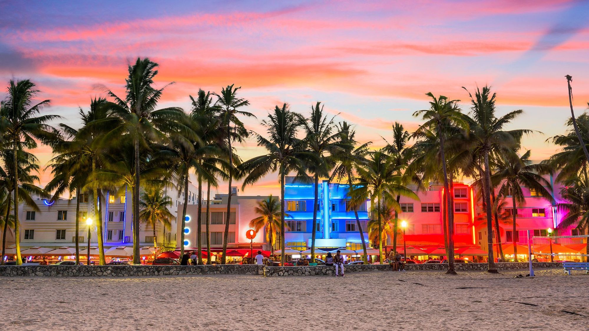 Ocean Drive i Miami Beach sett fra stranden, opplyst av fargerike neonskilt, med palmer mellom veien og sanden.