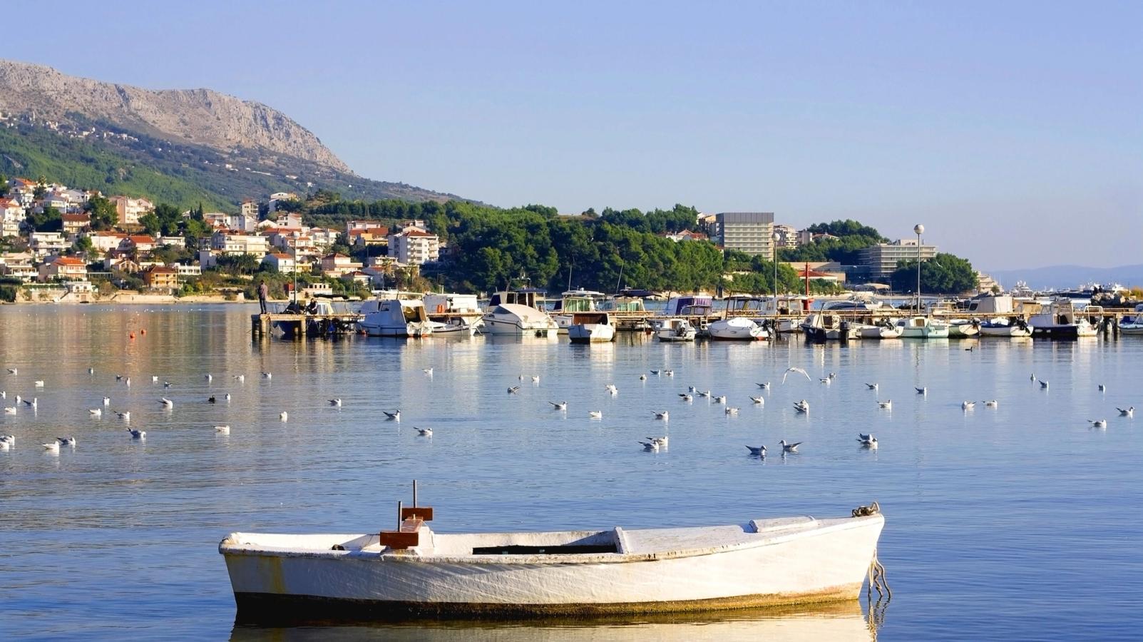 En lille hvid båd flyder stille på vandet i en bugt omgivet af måger. I baggrunden ses en marina med mange både, en kystby med røde tegltage, grønne træer og bjerge i horisonten under en klar blå himmel.