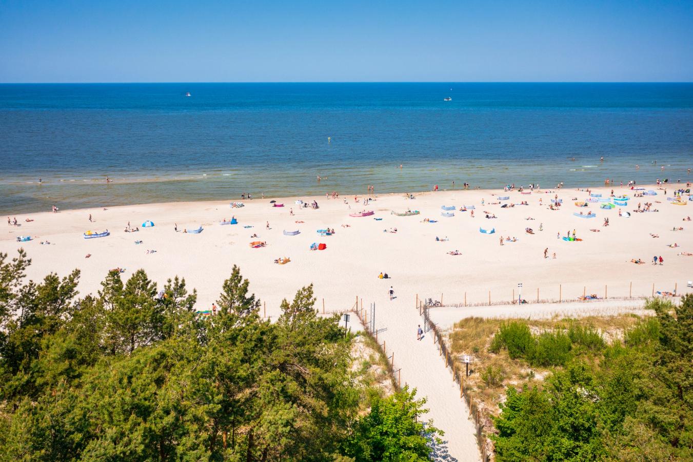 Bilden visar stranden vid Leba, längs Östersjön. I mitten den långa sandstranden med badgäster och parasoller. Bilden slutar med det stilla och blå vattnet i Östersjön.