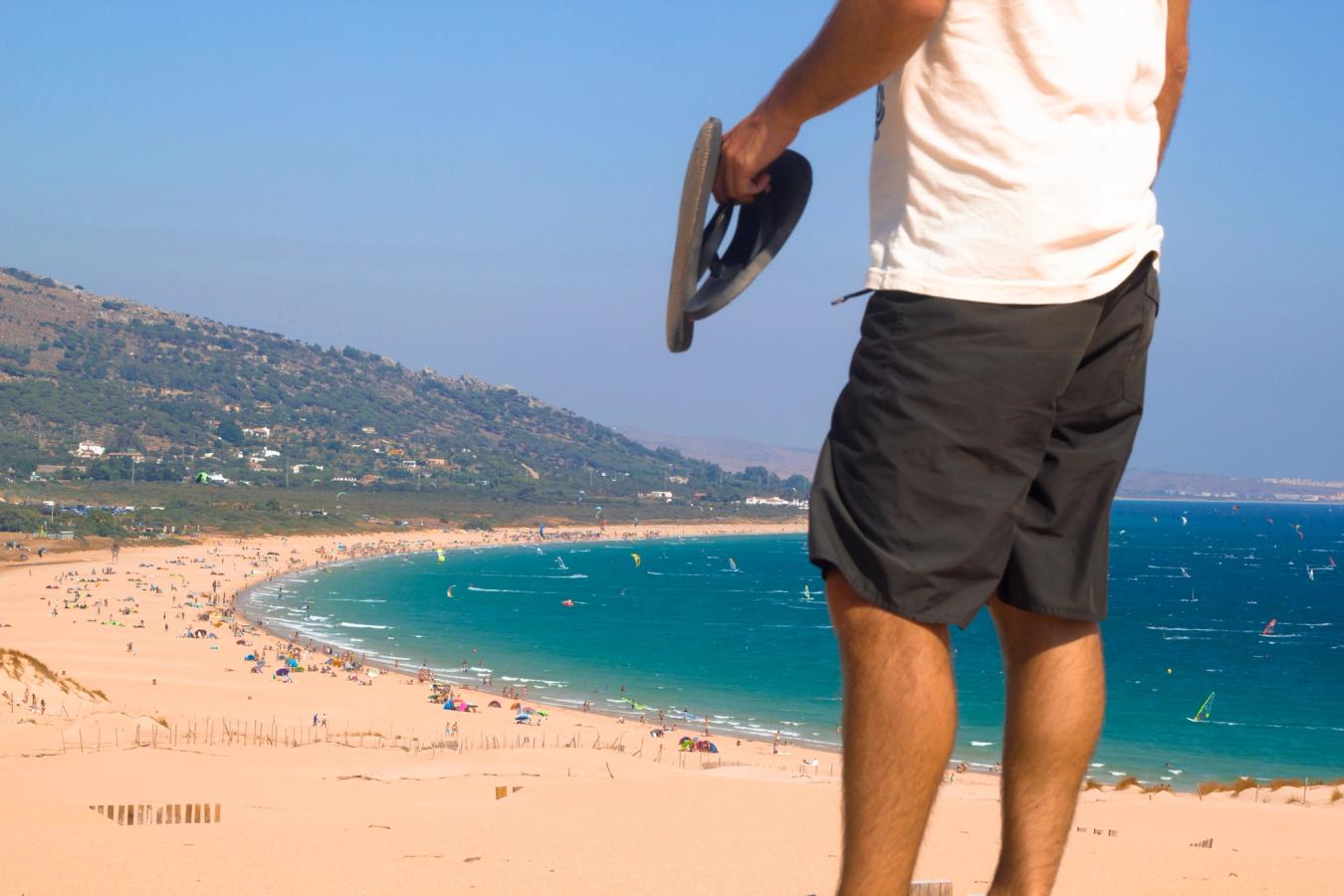 En mann ser utover stranden Playa de Bolonia fra en høyde. Stranden og vannet er fullt med surfere og kitere