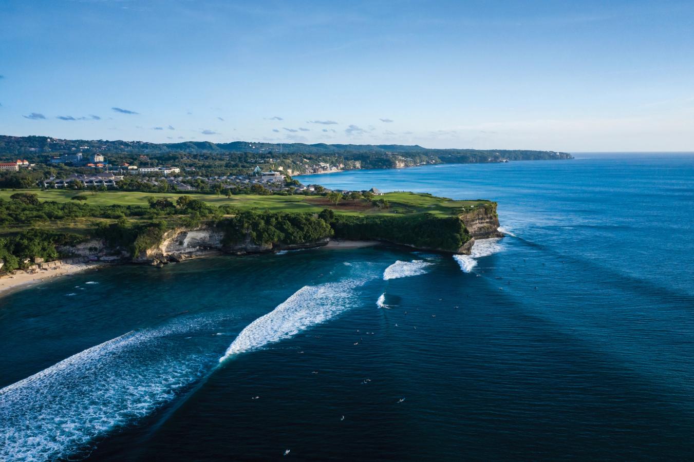 Balangan Beach, hvor store bølger ruller inn mot klippene. Man kan ane en liten strand, og ellers strekker landskapet seg med sin grønne vegetasjon inn mot byen som kan skimtes i bakgrunnen.