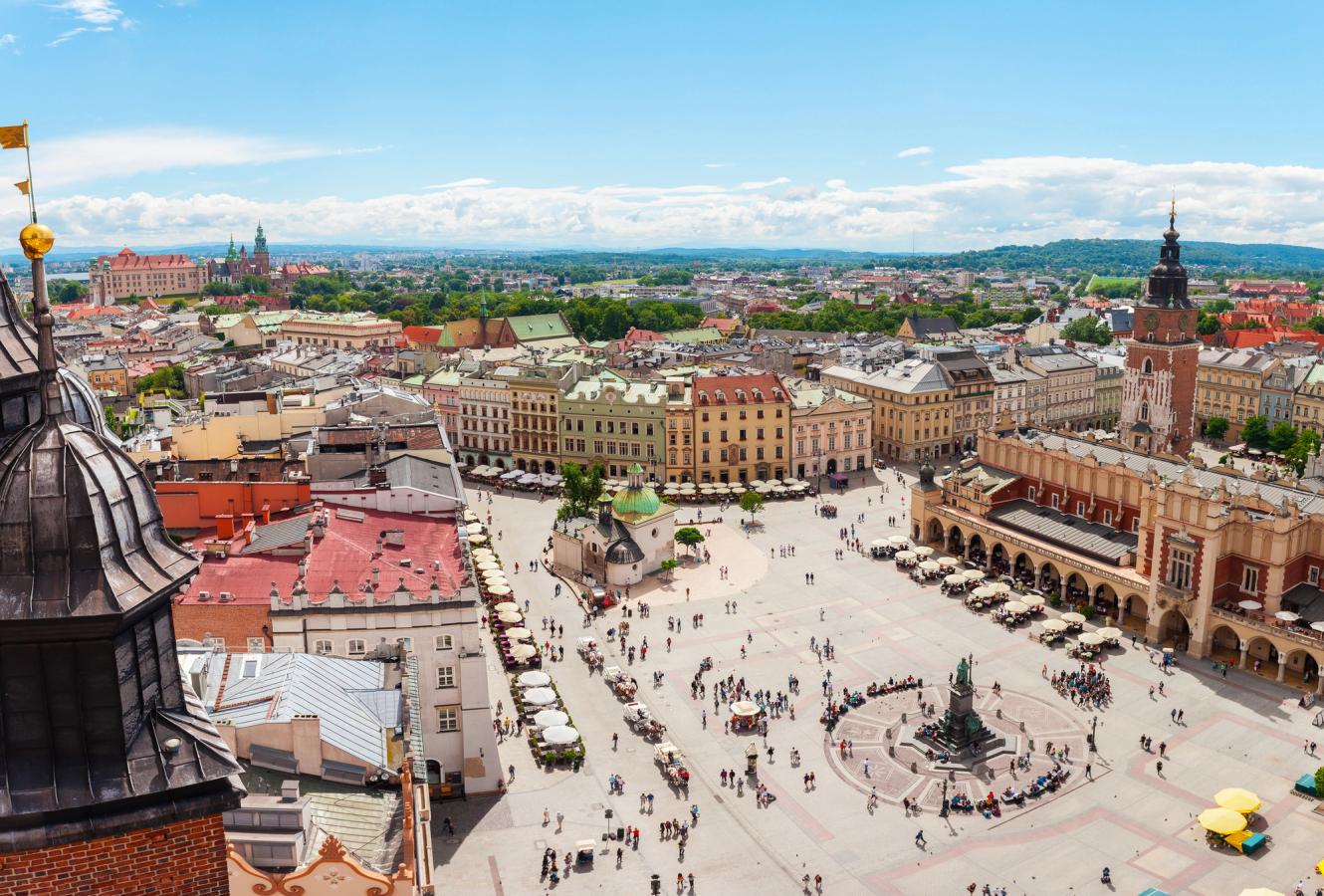 Bilden visar Krakows centrala torg och Kraków Cloth Hall (Sukiennice). I mitten av bilden finns en större öppen plats med ett konstverk i mitten. Torget är fullt av människor. På höger sida kan man se den låga och äldre byggnaden Kraków Cloth Hall. På vänster sida finns en rad med trottoarkaféer och stadsbebyggelse. I slutet av bilden ser man de karakteristiska byggnaderna i Gamla stan