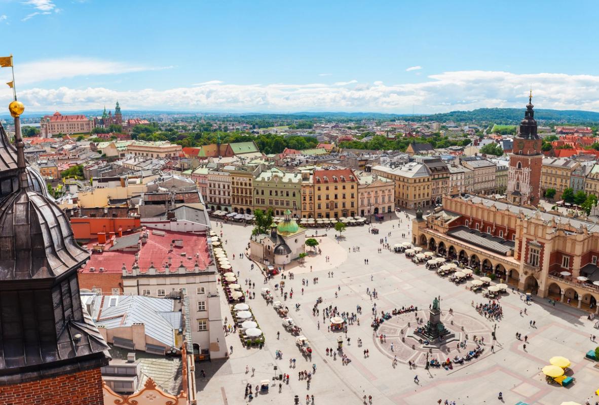 Bildet viser Krakow sitt sentrale torg og Kraków Cloth Hall (Sukiennice). I midten av bildet en større og åpen plass med et kunstverk i midten. Torget har en masse mennesker. På høyre side kan man se den lave og eldre bygningen Kraków Cloth Hall. På venstre side en rekke med fortauskafeer og bybebyggelse. Til slutt i bildet ser man de karakteristiske bygningene i gamlebyen.
