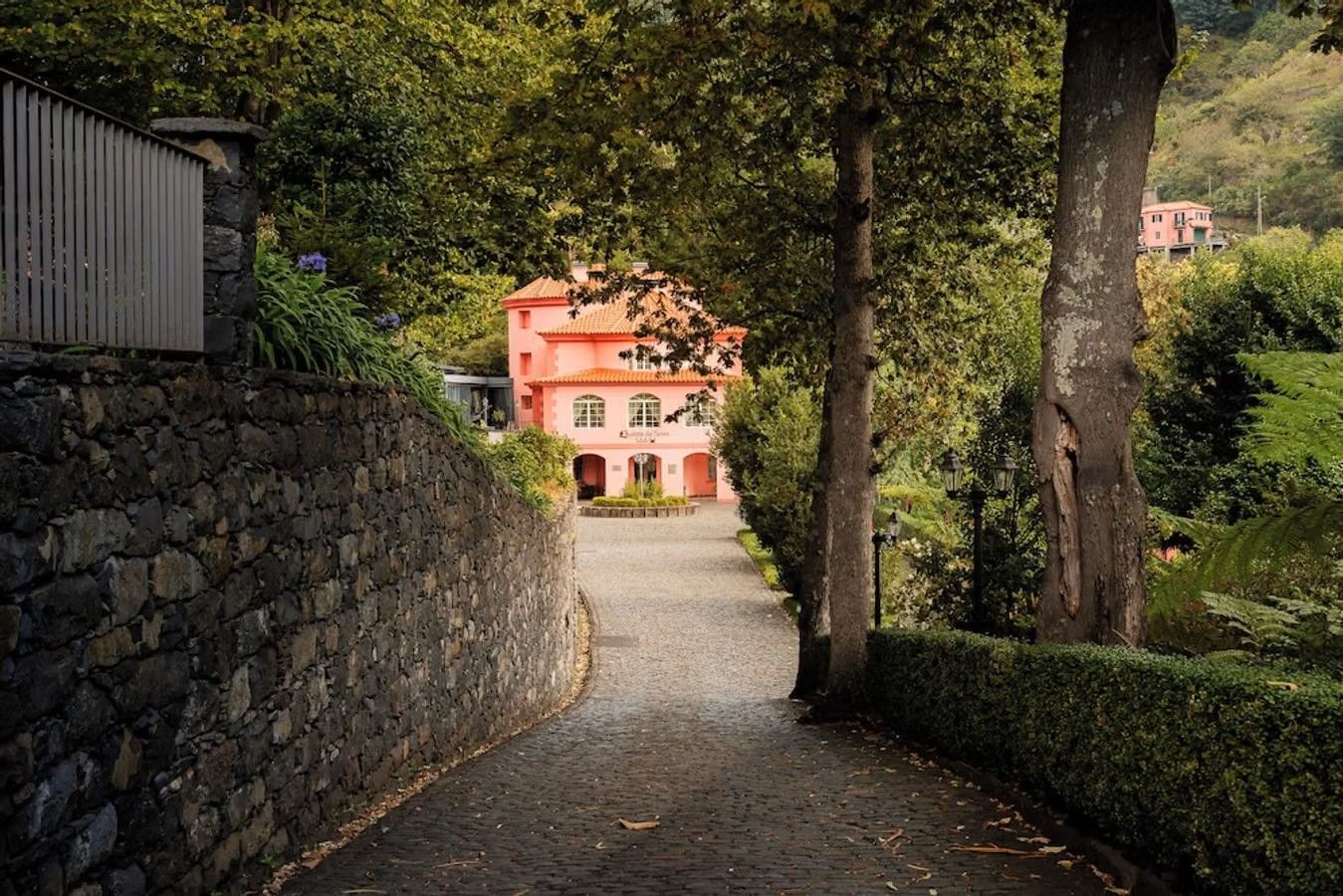 En smal brostensvej med en lav stenmur på hver side fører ind mod et tre-etagers murstenshus i rosa, som er Hotel Quinta Da Serra på Madeira.