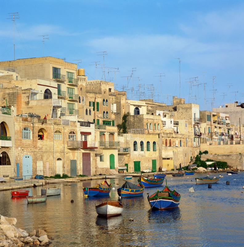Små fiskerbåde i havnen ved St. Julians på Malta. I baggrunden ses små lave huse med grønne skodder for vinduerne.