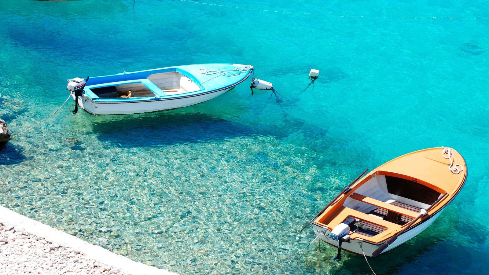 Två små motorbåtar ligger för ankar vid en stenstrand med kristallklart vatten.