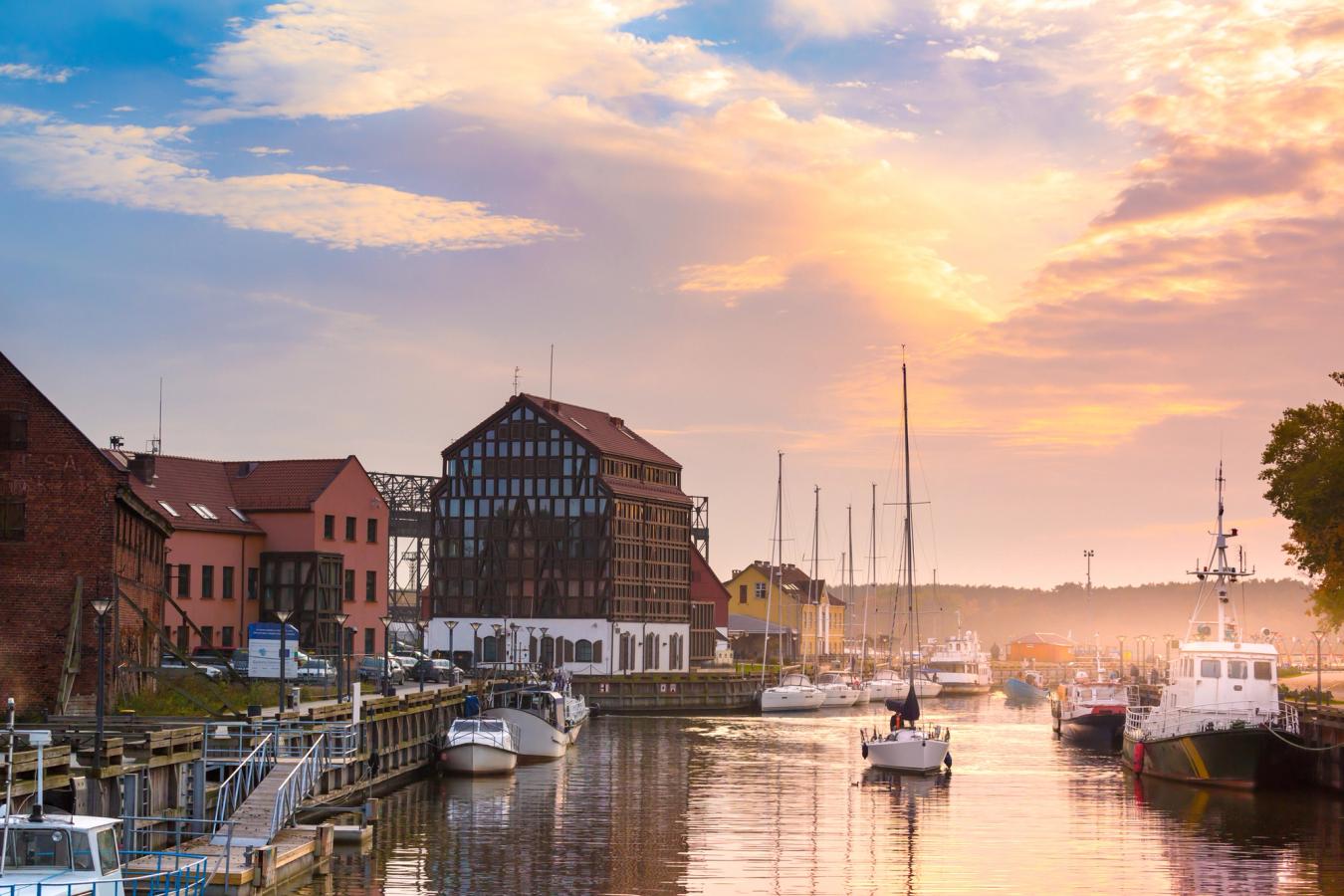 Bilden visar den mysiga hamnen i Klaipeda med små båtar som ligger vid kajen och några i vattnet. Korsvirkeshus på land