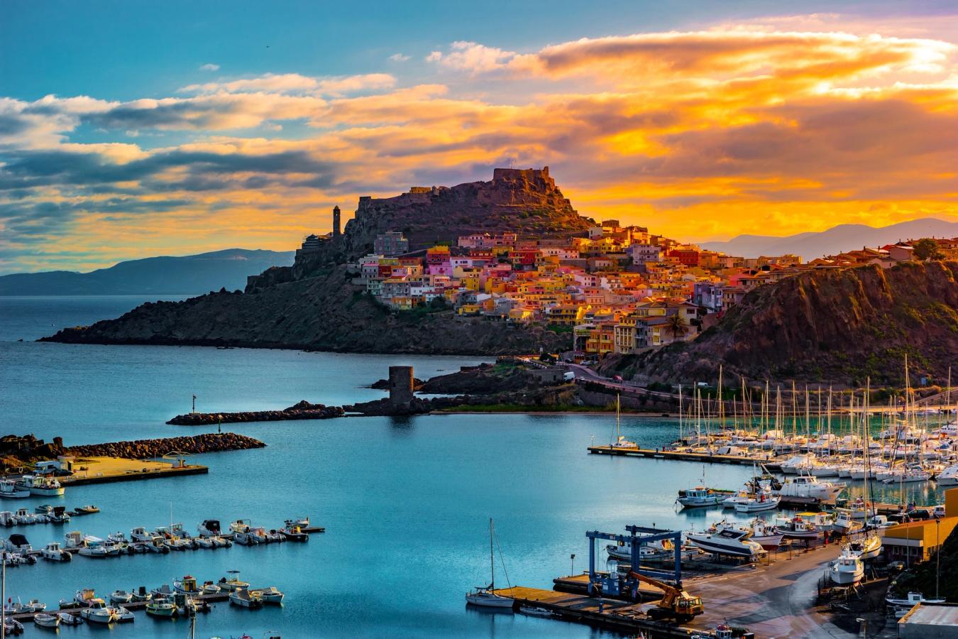 Bilden visar byn Castelsardo på Sardinien i solnedgången. Hamnområdet som övergår i större klippor där byn klamrar sig fast under en rödaktig himmel.