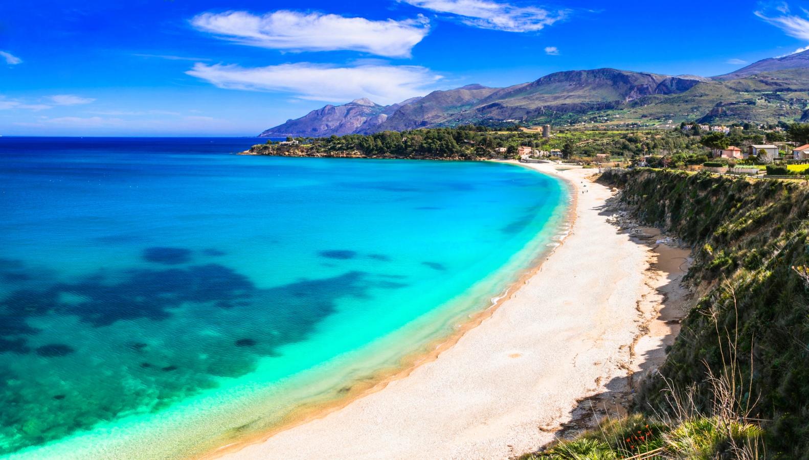 Bilden visar stranden Scopello Beach, precis vid Palermo på Sicilien i Italien. Stranden sträcker sig som en vik mot det turkosa och stilla Medelhavet. Bakom stranden, till höger, ligger ett frodigt landskap som övergår i en bergformation. Spridda moln på himlen