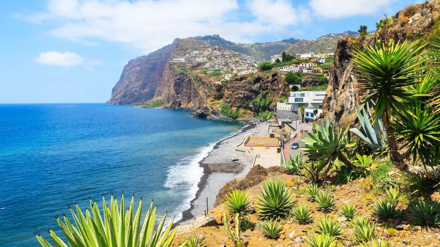 Udsigt over kystlinjen hvor små grønne kaktuslignende planter står på en skråning. Nedenfor ligger en klippestrand og en promenade. Atlanterhavet slår ind mod kysten og de stejle klipper hvor hvidmalede huse ligger spredt op ad bjergsiden.
