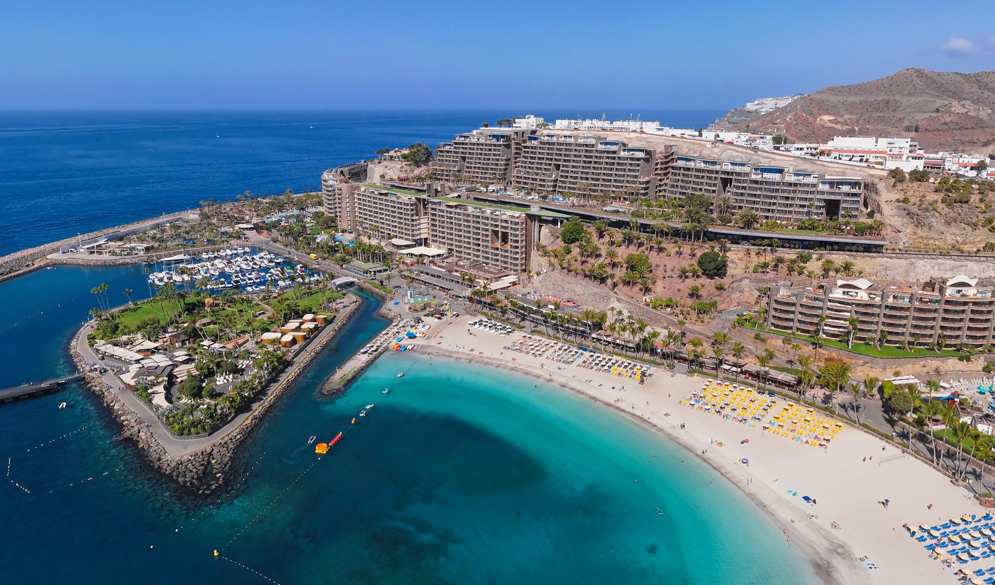 Her fra Playa Anfi del Mar i Puerto Rico på Gran Canaria. Stranden er sett ovenifra og viser en bred sandstrand med parasoller og solsenger. Til venstre er det bygget en marina på innsiden av en molo. I bakgrunnen av stranden ligger hotellene opp langs en åskam