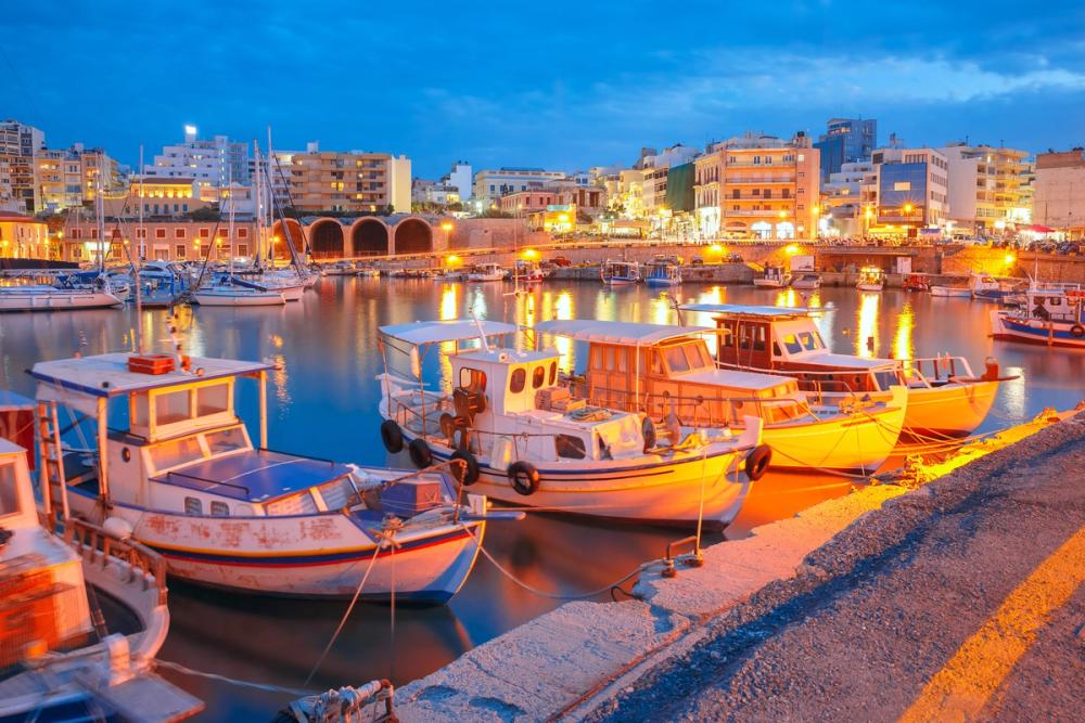 Bildet viser den gamle havna i Heraklion på Kreta. Fremst i bildet ligger små fiskebåter fortøyd, mens i bakgrunnen ser vi en kveldsopplyst havnepromenade med mennesker og turister. Bakenfor promenaden ligger en lavere 4 etasjers bebyggelse med en mørkere blå himmel