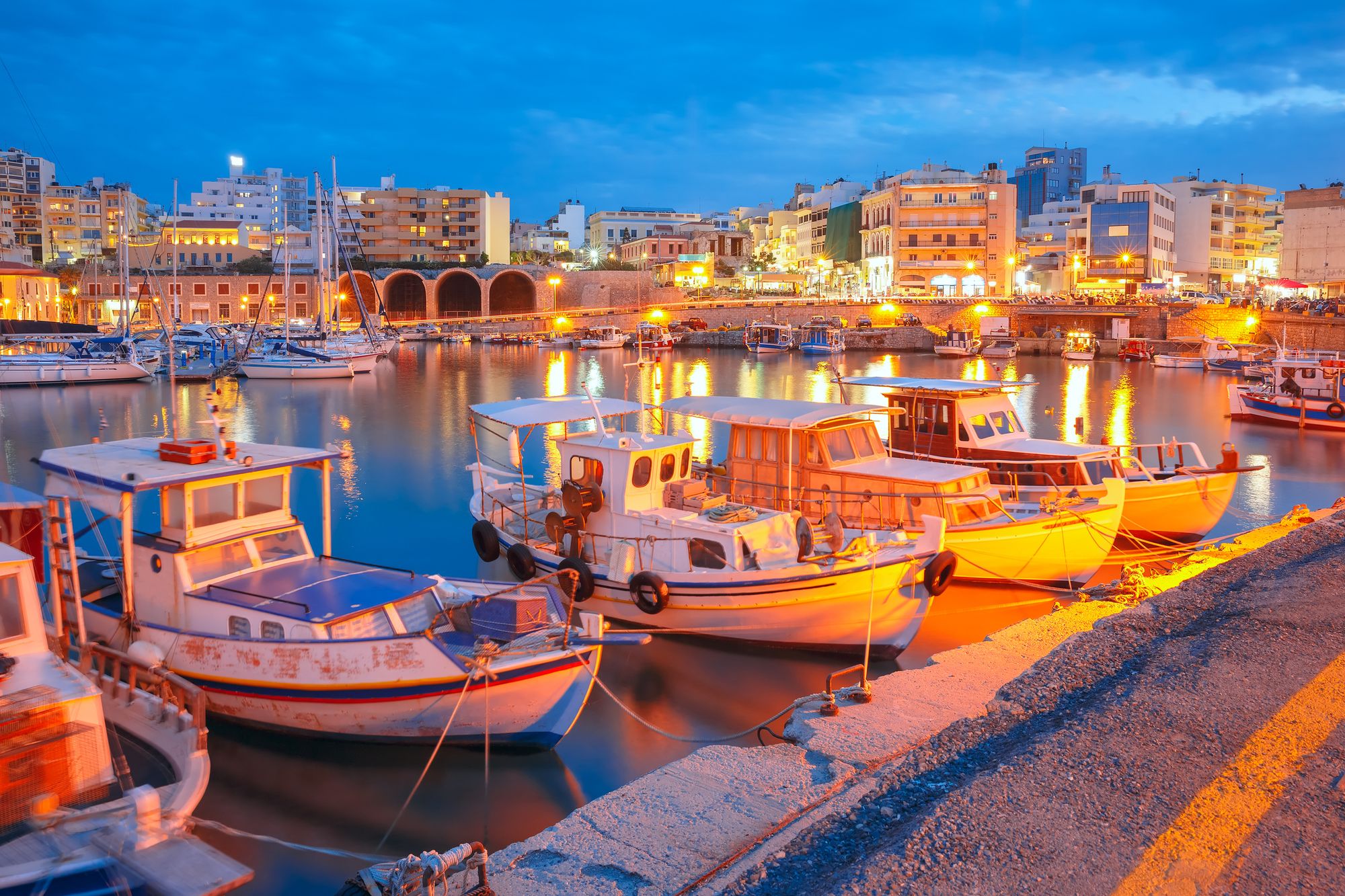 Bildet viser den gamle havna i Heraklion på Kreta. Fremst i bildet ligger små fiskebåter fortøyd, mens i bakgrunnen ser vi en kveldsopplyst havnepromenade med mennesker og turister. Bakenfor promenaden ligger en lavere 4 etasjers bebyggelse med en mørkere blå himmel