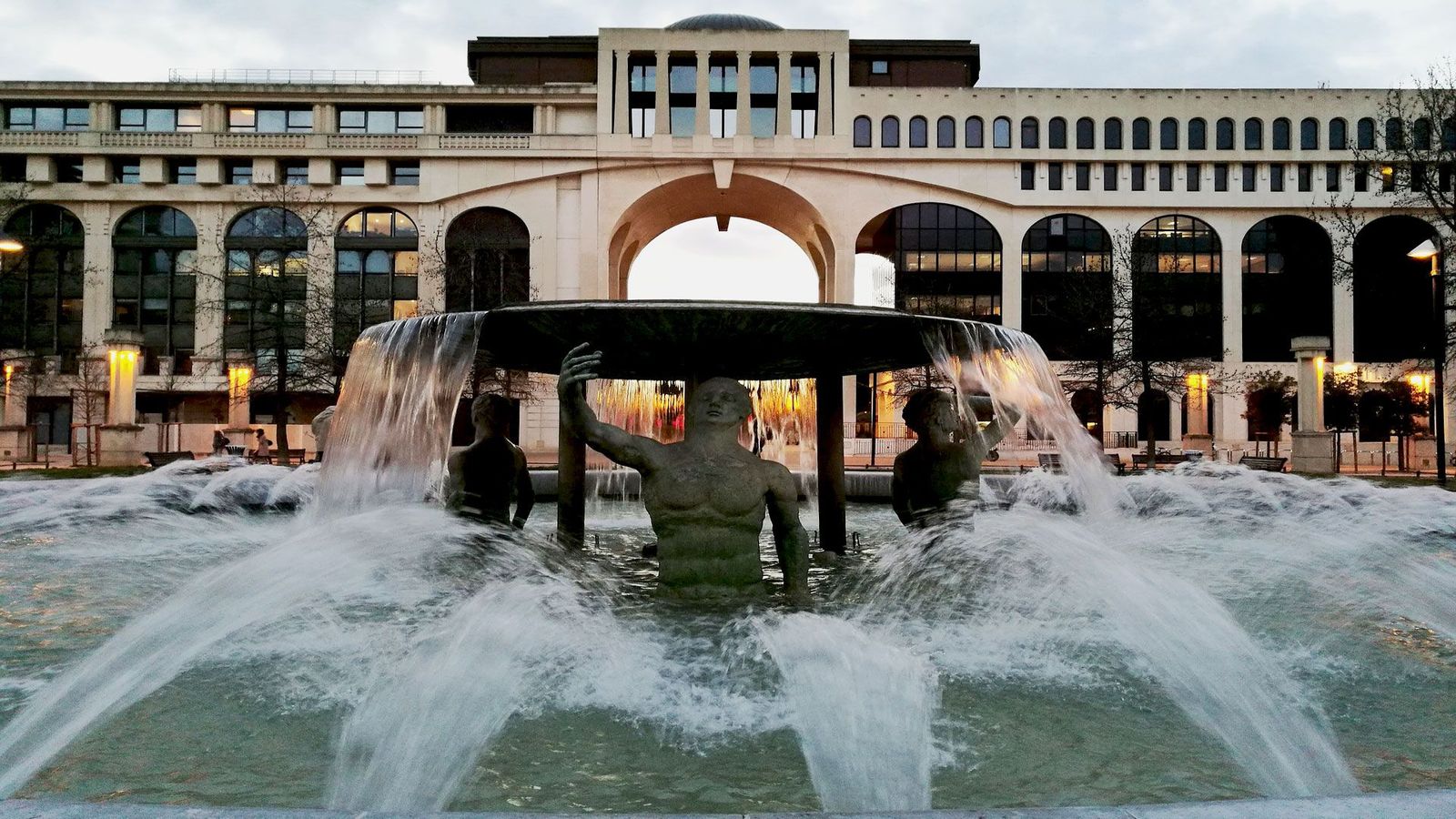 Et nærbillede af et springvand, hvor skulpturer af mennesker står i en cirkel og bærer en tynd, rund plade. I baggrunden ses en bygning med et moderne islæt.