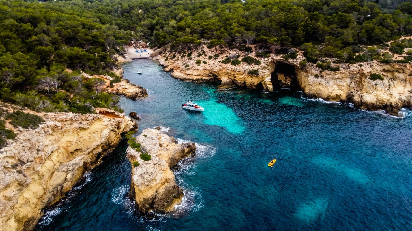 Billedet, der er taget med en drone, viser bugten Cala Falco. Den lille bugt er omgivet af lavere klippevægge og ender i en smal sandstrand. Både klipperne og stranden er omgivet af frodig, grøn skov.