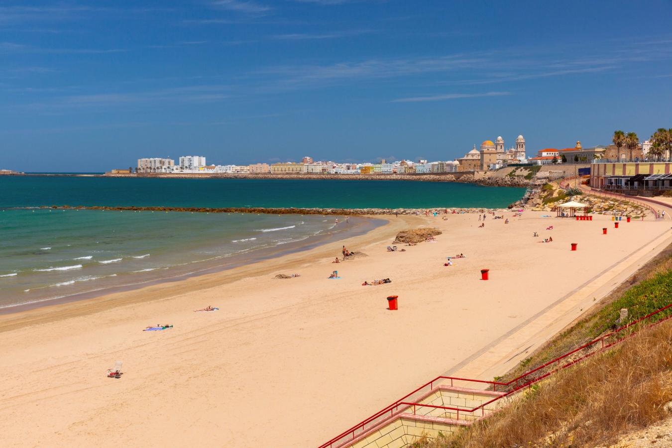 En lang og bred sandstrand strekker seg inn mot byen Cadiz. Havet er grønt, og bølgene har små hvite skumtopper.