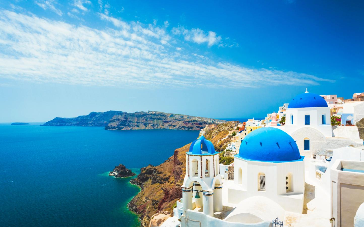Bilden visar en del av staden Oia på Santorini i Grekland. Till höger vitkalkade byggnader med karakteristiska koboltblå och välvda tak. Till vänster ser vi branta klippväggar som går ner i det blå Medelhavet. Spridda moln på himlen