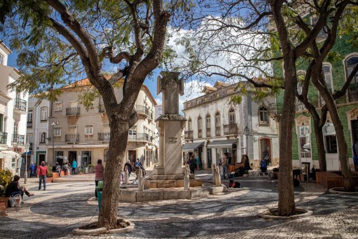 Ett litet torg i Lagos med ett monument i mitten, omgivet av några träd. På torget finns flera människor, och det är omgivet av låga hus med små, utstickande balkonger. Bottenvåningen i husen är inriktad på caféer, med små markiser som hänger ut över trottoaren. Himlen är blå med några lågt hängande moln.