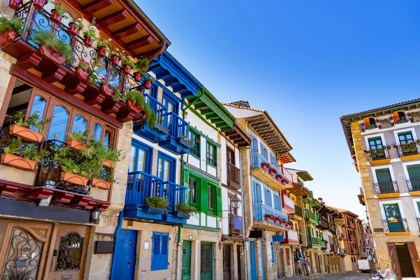 En smal gata med tätt liggande hus i Hondarribia. Flera av husen har balkonger i olika färger och små balkonglådor med blommor. Över hustaken syns himlen som är helt klar och blå.