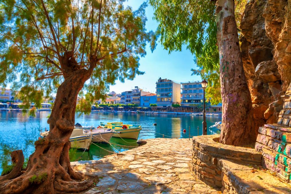 Bildet viser den lille innsjøen Voulismeni Lake i byen Agios Nikolaos på Kreta. Fremst i bildet ser vi en promenade langs vannet belagt med steinheller. På hver side av promenaden vokser det lokaltypiske trær. I vannet er det tre små fiskebåter fortøyd. Innover i bildet ser vi det blikkstille vannet og lengst inn kan vi se restauranter langs promenaden og litt høyere bebyggelse bakenfor. Alt med en skyfri og blå himmel.