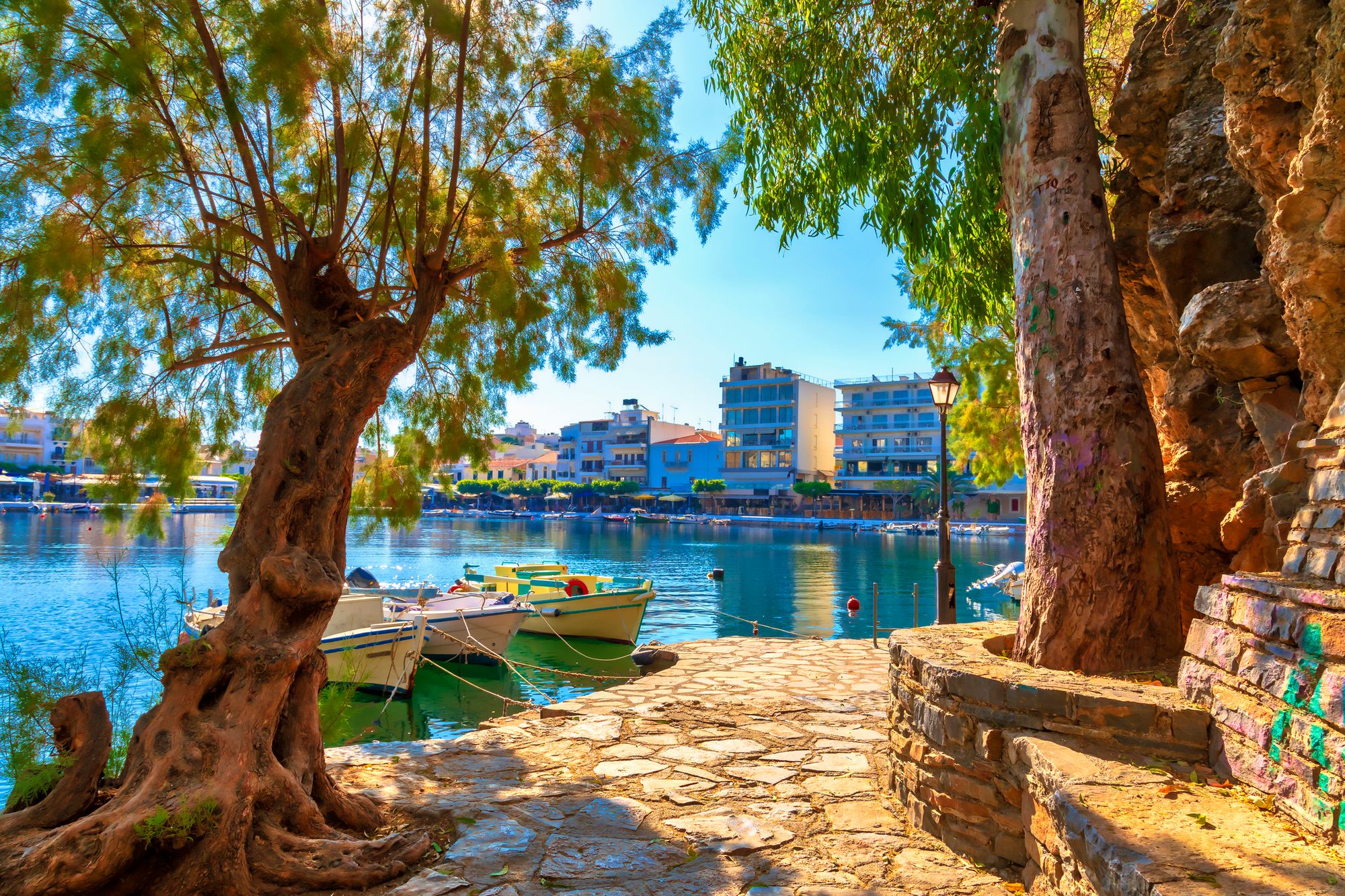 Bildet viser den lille innsjøen Voulismeni Lake i byen Agios Nikolaos på Kreta. Fremst i bildet ser vi en promenade langs vannet belagt med steinheller. På hver side av promenaden vokser det lokaltypiske trær. I vannet er det tre små fiskebåter fortøyd. Innover i bildet ser vi det blikkstille vannet og lengst inn kan vi se restauranter langs promenaden og litt høyere bebyggelse bakenfor. Alt med en skyfri og blå himmel.