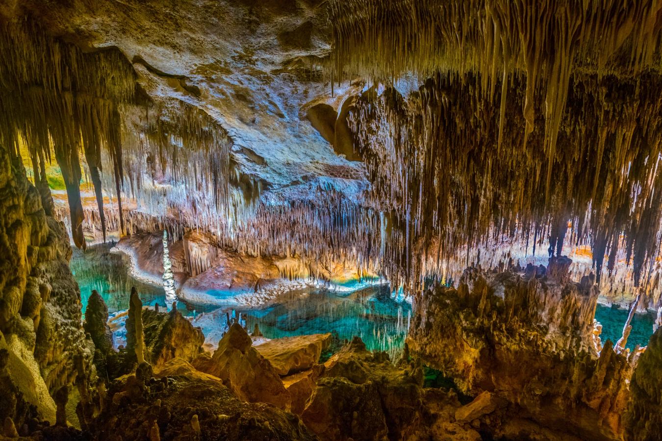 Billedet viser indersiden af Dragon Cave på Mallorca. Fra loftet i grotterne kan man se et væld af stalaktitter/drypsten "vokse" både fra loftet og ned og op fra "gulvet". I midten af billedet kan man se den turkisblå sø, der har dannet sig i grotterne.