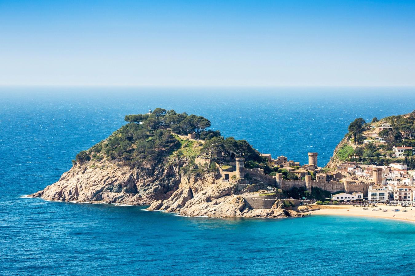 Bildet viser murene til Vila Vella på Tossa de Mar Beach langs Costa Brava. Man ser bystranden nederst i høyre hjørnet, med bymurene til venstre rundt en klippehøyde. Stille vann og alt under en skyfri himmel.