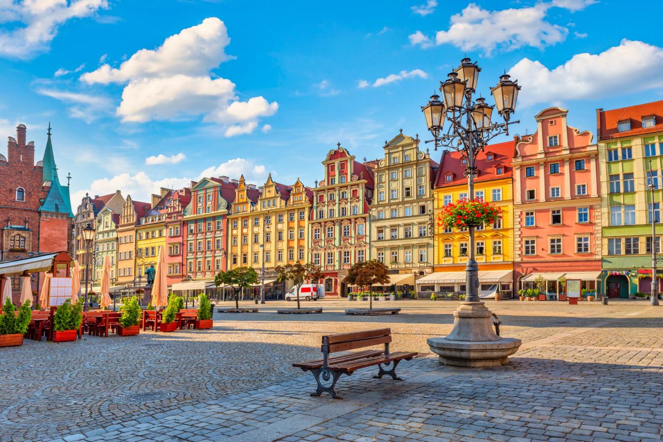 Bildet er fra Wrocławski Rynek i gamlebyen i Wroclaw. Vi ser torget i midten som er brosteinsbelagt. Langsetter torget ser vi de smale byggverkene i ulike pastellfarger. Enkelte fortauskafeer langs bygningene. Spredte skyer på himmelen.
