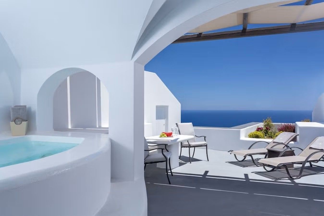 Bilden visar det helvita och kalkade rummet du får på hotellet Amber Light Villas på Santorini. Man kan se delar av en vitklädd jacuzzi till vänster och öppningen ut mot den privata terrassen med solstolar och kafébord. Utsikten visar en kristallblå himmel och ett hav som nästan går i ett.