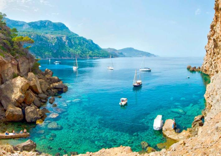 Bildet viser en av Mallorca sine mange små bukter med krystallklart vann. Her med klippeformasjoner rundt bukten, båter på stille vann og med frodig og grønt landskap i bakgrunnen.