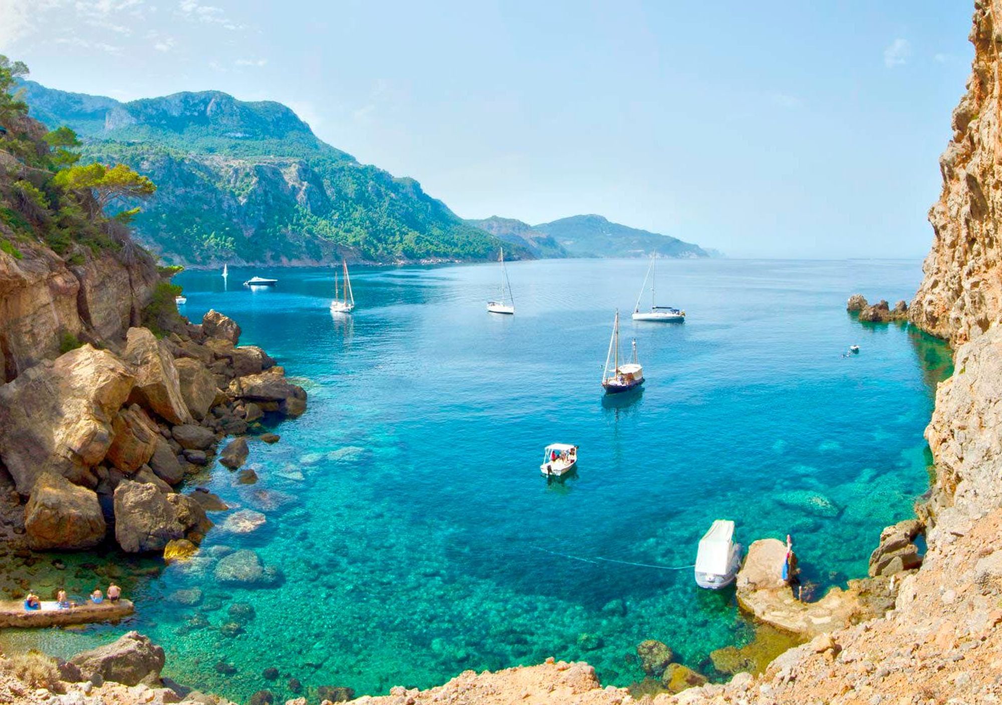 Billedet viser en af Mallorcas mange små bugter med krystalklart vand. Her kan man se klippeformationer rundt om bugten, både på roligt vand og et frodigt grønt landskab i baggrunden.