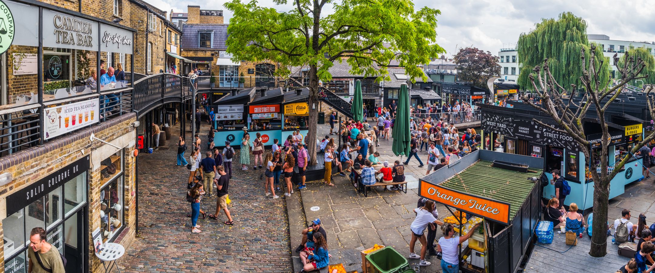 Billedet viser en aktiv dag på Camden Market. Her med et åbent "torv" med et par grønne træer, omgivet af toetagers bygninger i kombination med mindre boder. Bygningerne og boderne er en blanding af mad- og tøjsalg. Torvets midte er delvis fuld af mennesker, der shopper, spiser og vandrer rundt på torvet.