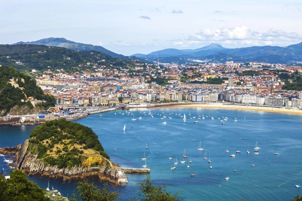 Oversiktsbilde over San Sebastian sett fra sjøsiden, med vannet fullt av ulike båter, bystranda i en bue og bybebyggelsen i bakkant. Lengst inn i bildet skogkledde fjellformasjoner.