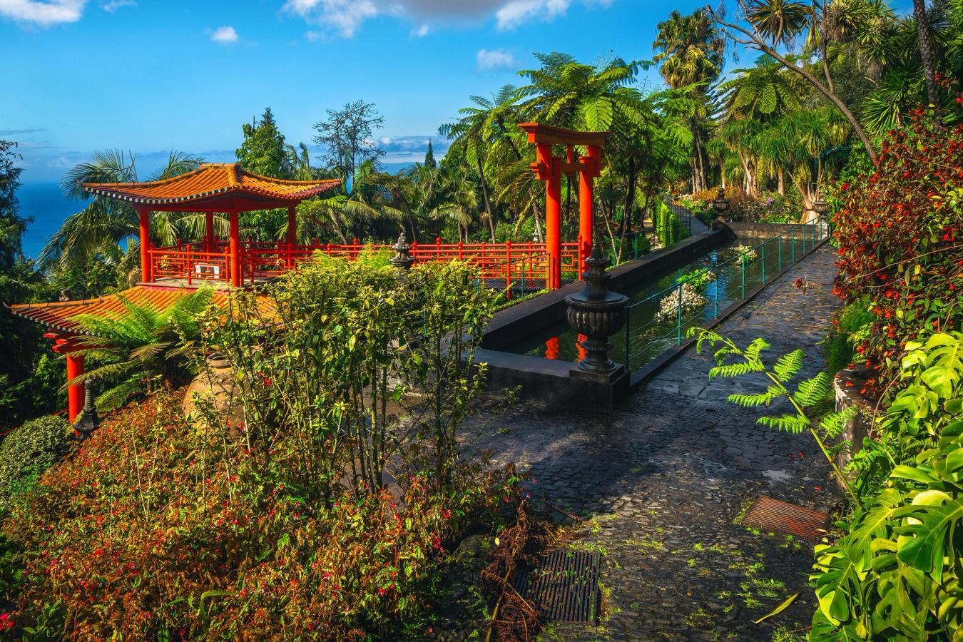 Bilden visar Monte Palace Tropical Gardens på Madeira. Ett frodigt trädgårdslandskap, japanskinspirerat och med en röd terrassliknande utsiktspunkt. Vid ingången ligger en mindre vattenspegel. Allt under en blå himmel.