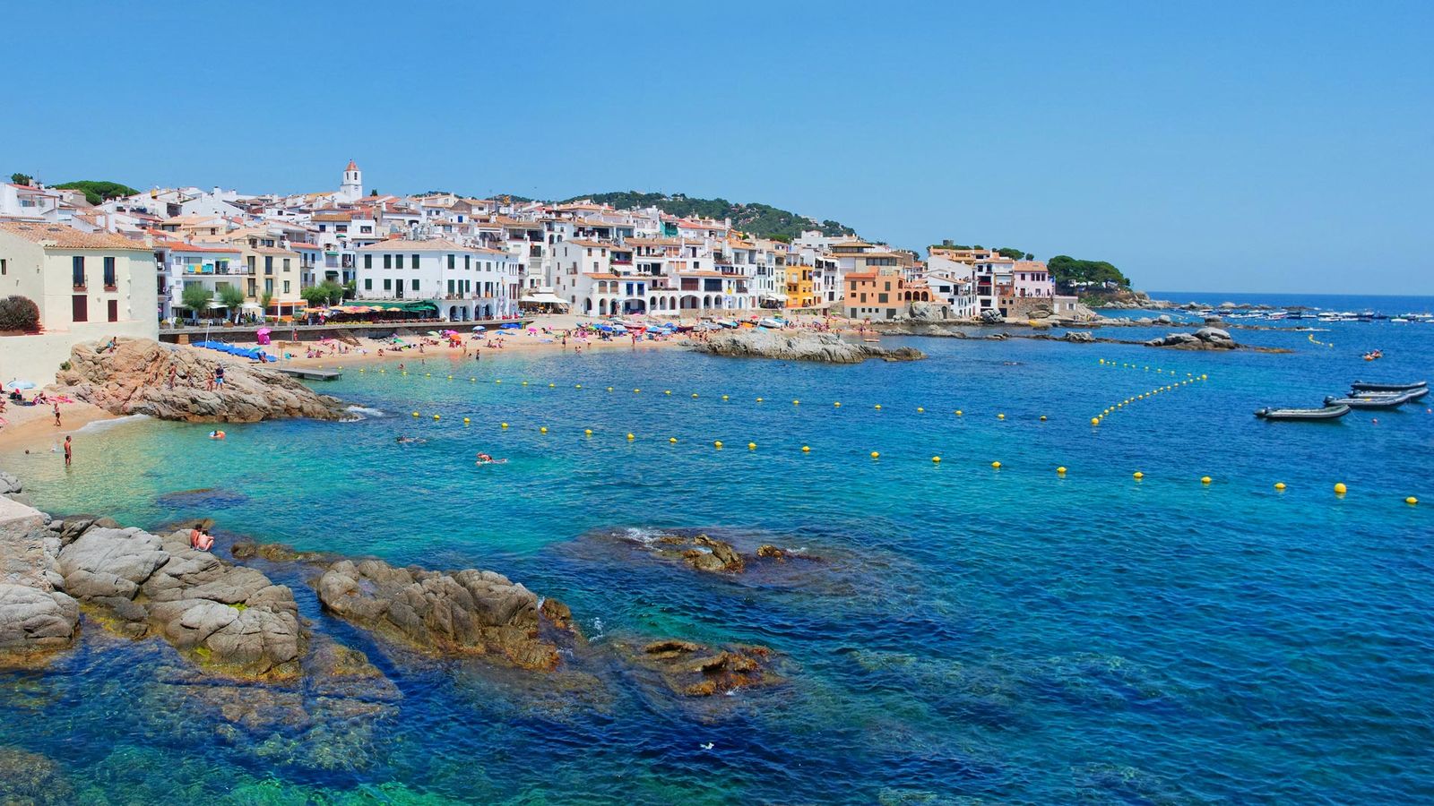 Små hus med röda tegeltak ligger alldeles vid det kristallklara havet. Framför bebyggelsen finns en liten sandstrand, medan det i vattnet finns flera stenar och små klippformationer. I horisonten skymtar det gröna landskapet.