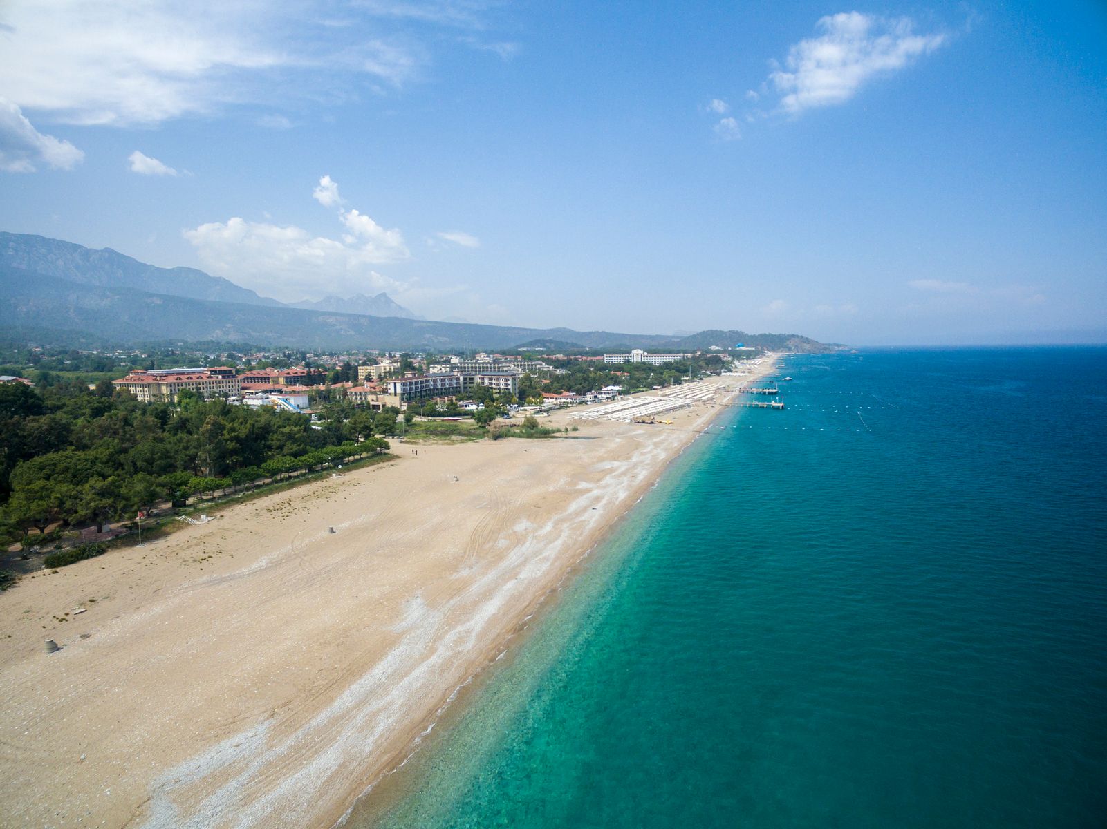 Utsikt over en langstrakt sandstrand med hvite parasoller. Bak stranden ligger bebyggelse, og bak dette reiser det seg flere fjell. Det turkisblå havet utenfor stranden er rolig, og himmelen er blå med noen få skyer.