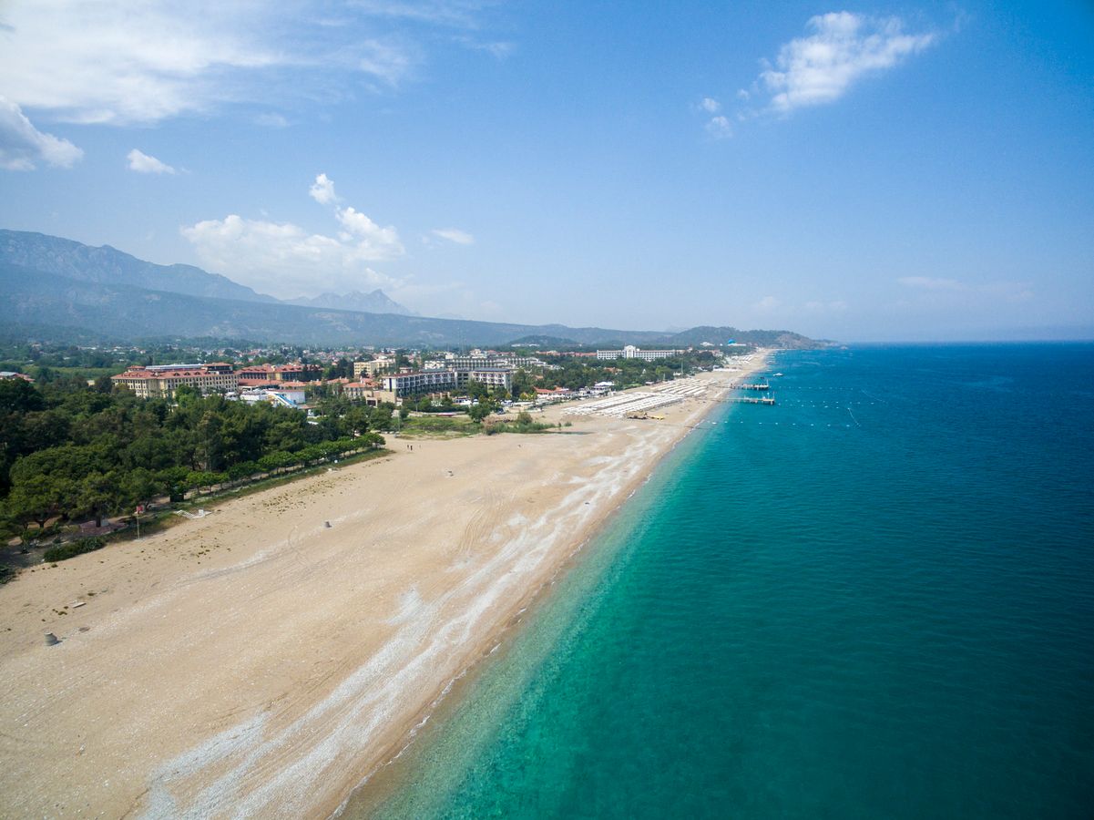 Udsigt over en langstrakt sandstrand med hvide parasoller. Bag stranden er der bebyggelse, og bag dette er der flere bjerge.  Det turkisblå hav ud for stranden er roligt, og himlen er blå med få skyer. 