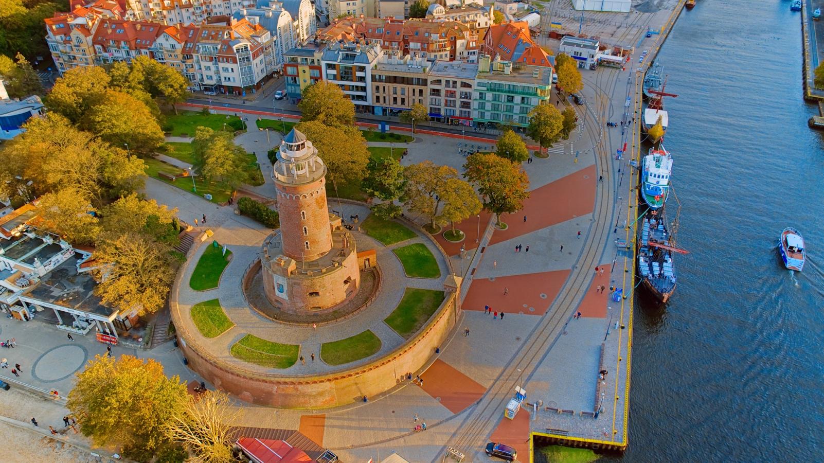 Billedet viser fyrtårnet i Kolobrzeg. Fyrtårnet er lavet af rødbrun mursten omgivet af grøn vegetation. I baggrunden ser vi den ældre bebyggelse med promenaden og både fortøjet til højre.