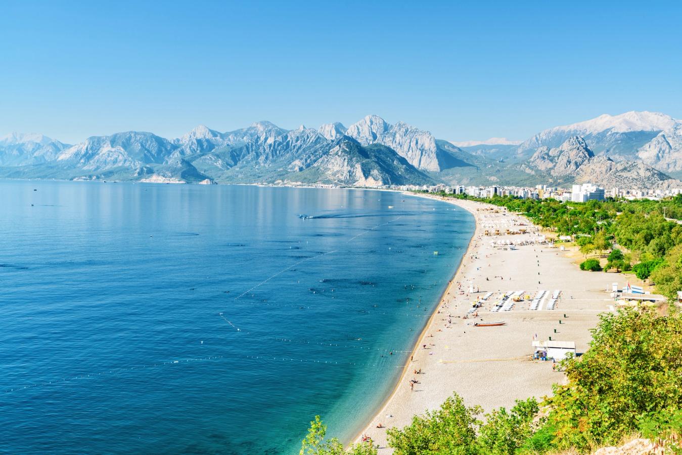 Billedet viser Konyaalti Beach midt i billedet, langs Antalyakysten med det blå Middelhav til venstre. Længst inde ser vi en større bjergkæde under en skyfri himmel.